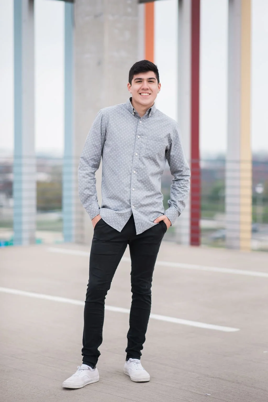 A young man standing outdoors on a rooftop with a city view, wearing a light gray button-up shirt with small white polka dots, black pants, and white sneakers, smiling with hands in pockets.