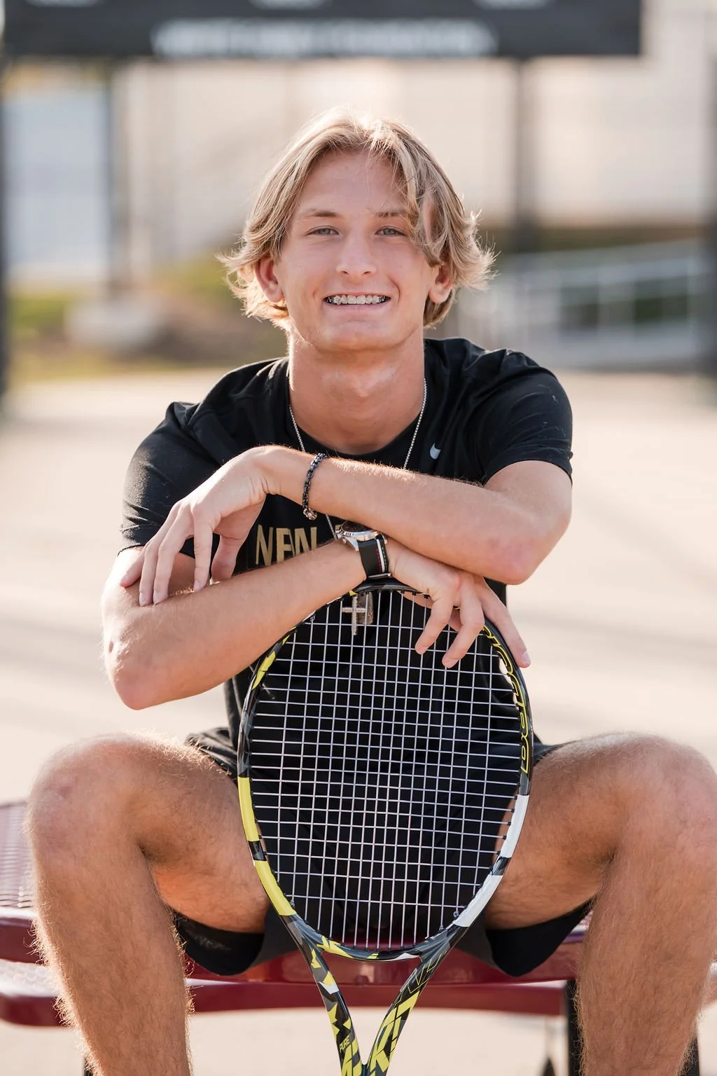 A young man with blonde hair sitting on a bench outdoors, holding a tennis racket across his lap. He is smiling and dressed in athletic clothing.