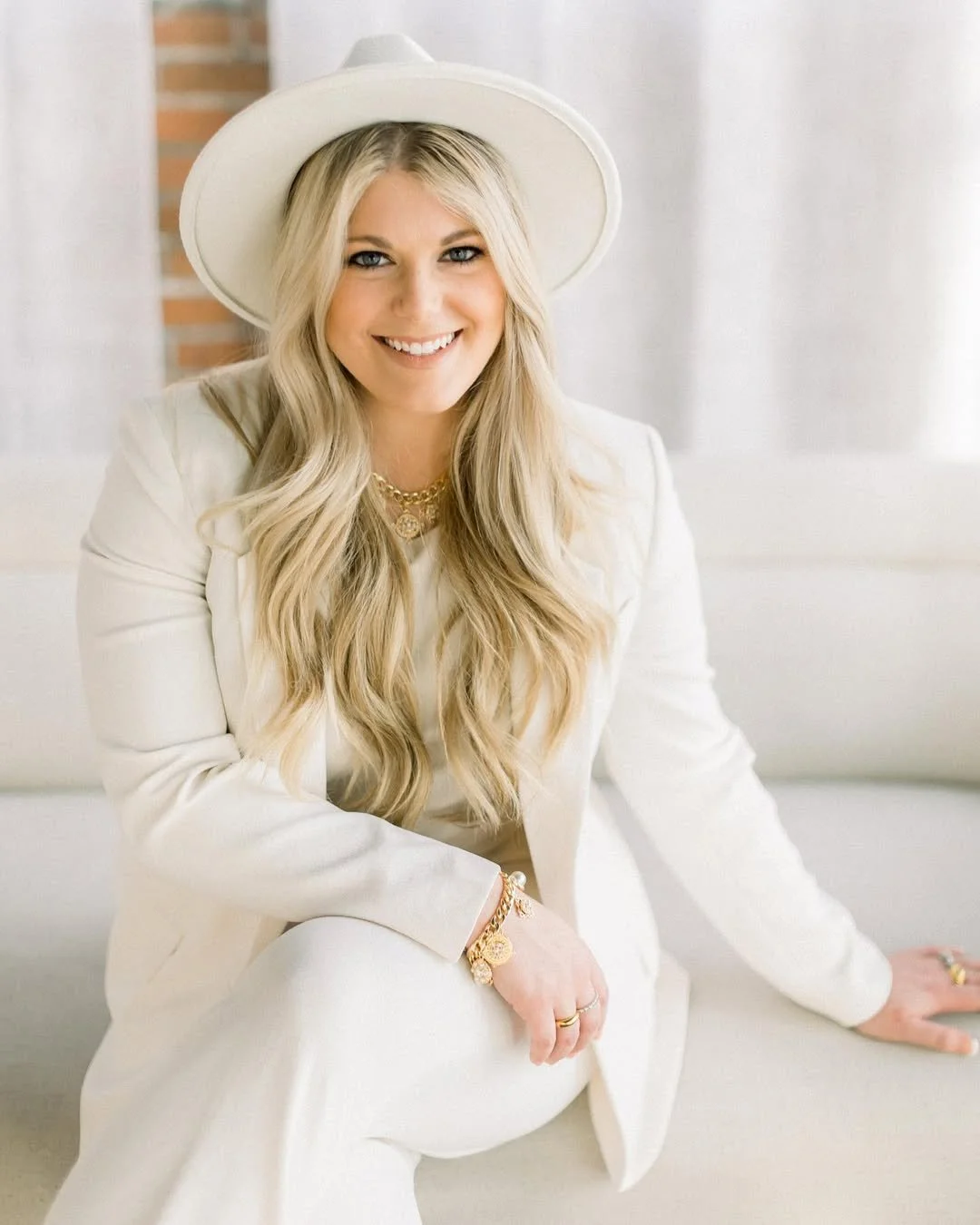 A smiling woman with long blonde hair, wearing a white hat and white suit, sitting on a white couch inside a bright room with a brick wall in the background.