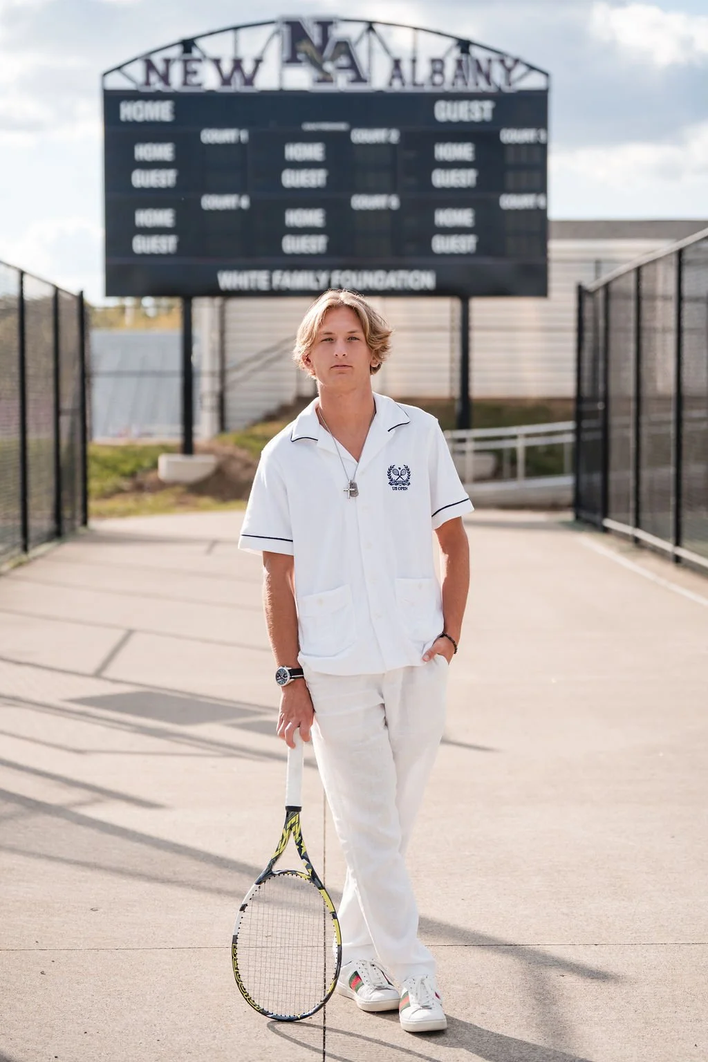 A young man standing on a tennis court holding a tennis racket, wearing a white tennis outfit with a logo, with a scoreboard and fence in the background.