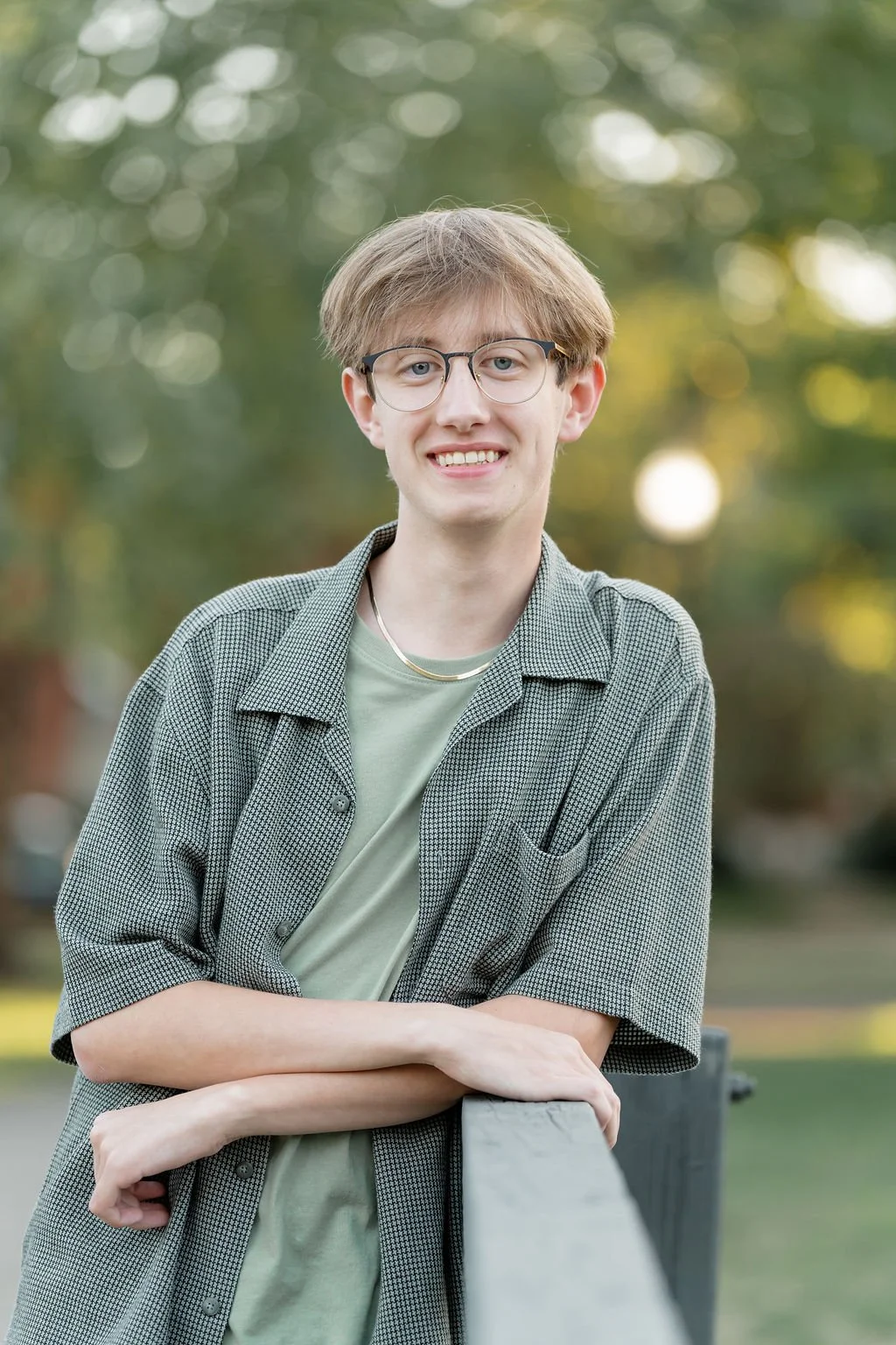 A young man with glasses and light brown hair, smiling, leaning on a gray railing outdoors with blurred trees and sunlight in the background.