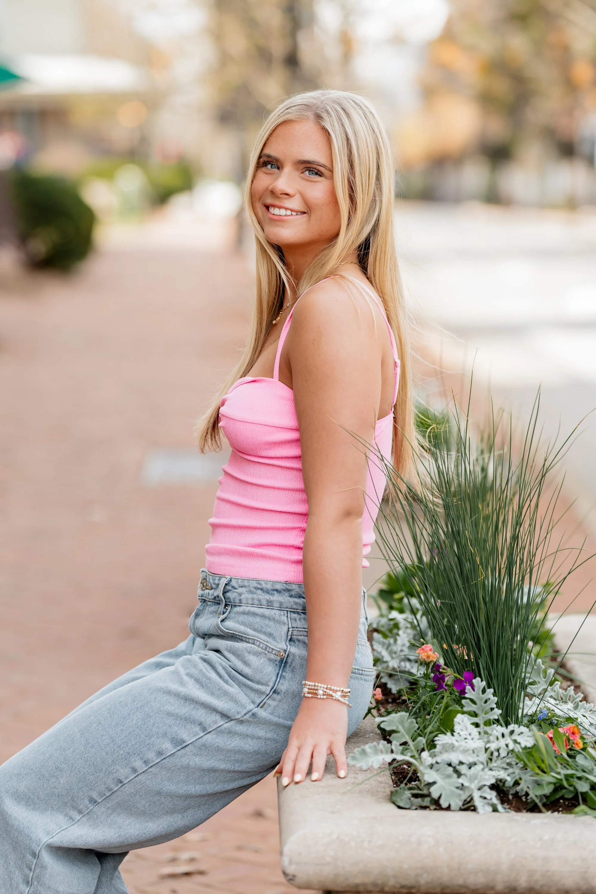 A young woman with long blonde hair smiling, sitting on a park bench next to a flower bed with green plants and colorful flowers, wearing a pink tank top and high-waisted jeans.