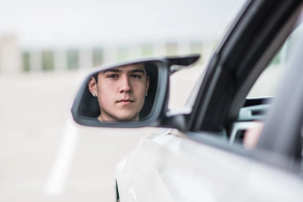 A young man with earrings looking at his reflection in a car's side mirror