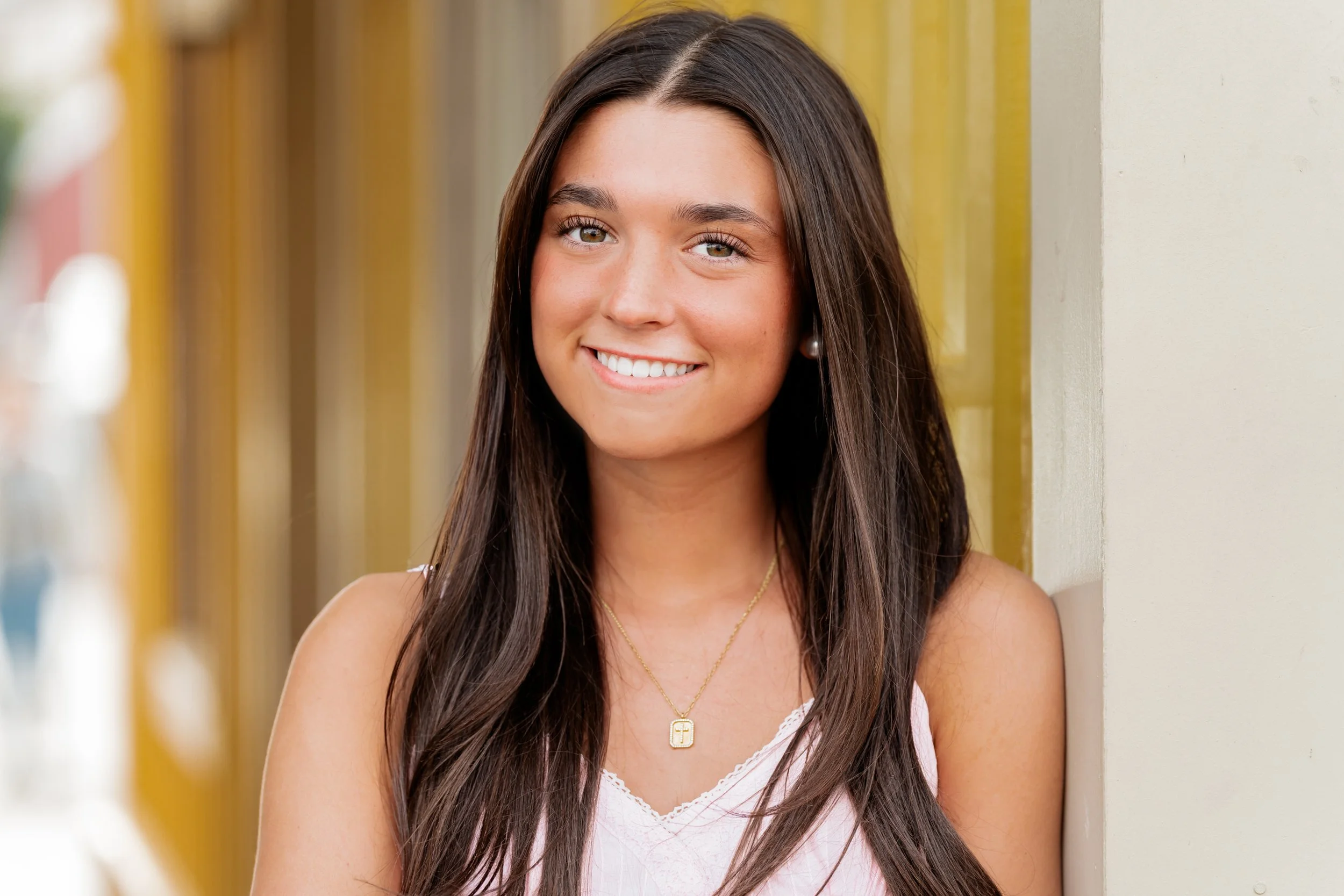 A young woman with long brown hair, smiling, wearing pearl earrings, a gold necklace, and a pink top, standing outdoors near a yellow wall.