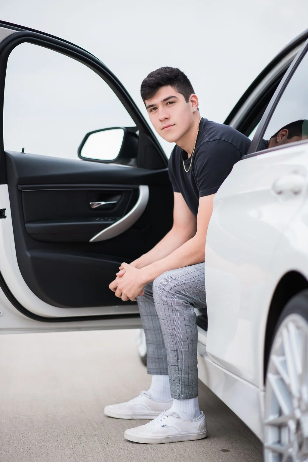 A young man with short dark hair, dressed in a black t-shirt, plaid gray pants, white sneakers, and jewelry, sitting in the open door of a white car with a gray interior against a plain, neutral background.