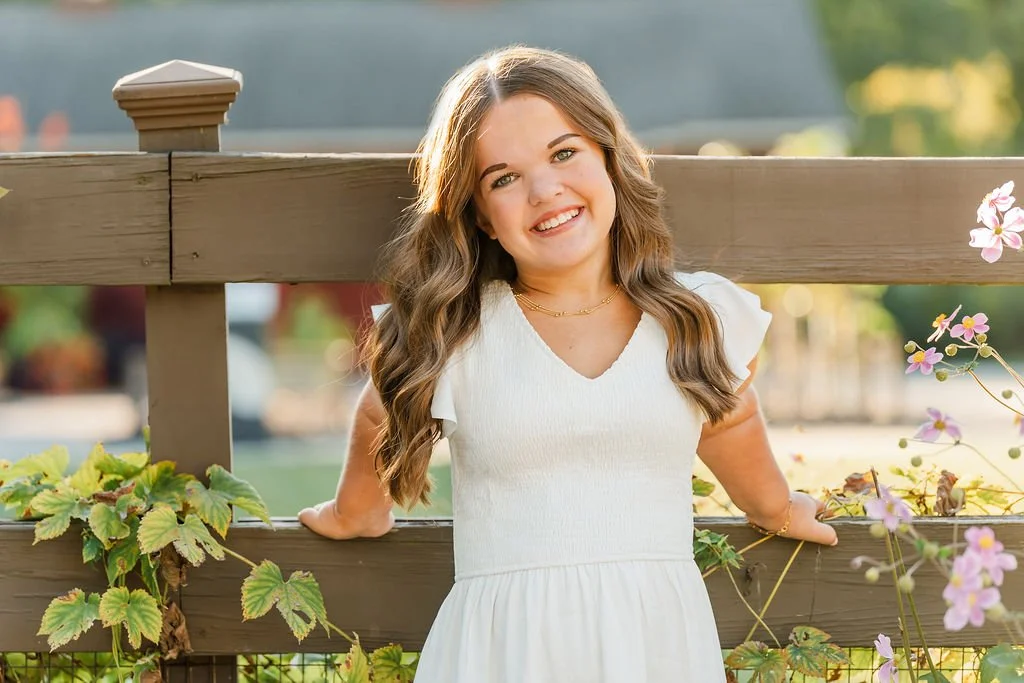 A young woman with long wavy brown hair, smiling, wearing a white dress and a gold necklace, leaning on a wooden fence with green and purple plants, in an outdoor setting with blurred trees and buildings in the background.