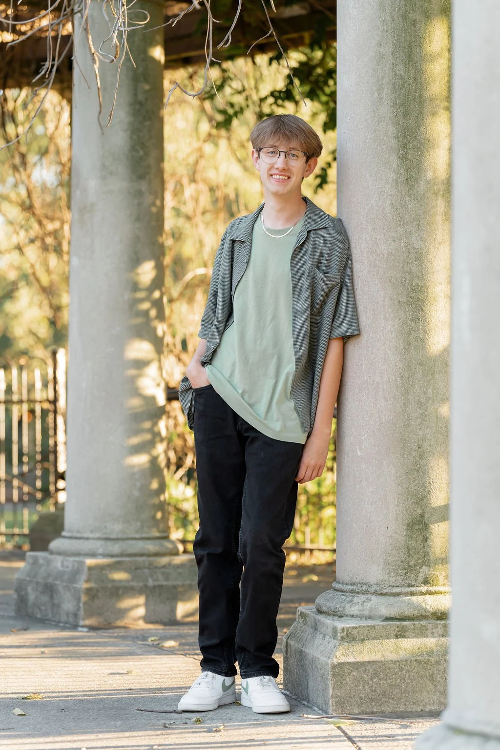 A young man with glasses and brown hair, wearing a light green t-shirt, black jeans, and a checkered short-sleeve shirt, is standing outdoors next to a large stone column, smiling at the camera.