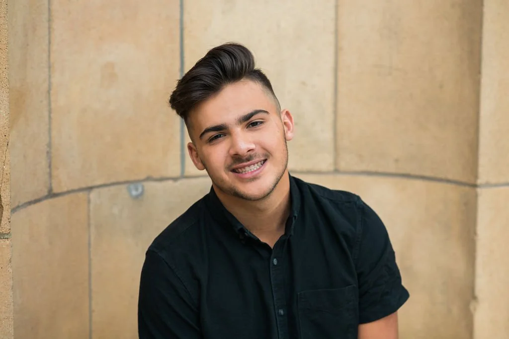 A young man with styled brown hair, fair skin, and a small beard, wearing a black button-up shirt, smiling and leaning against a beige stone wall.