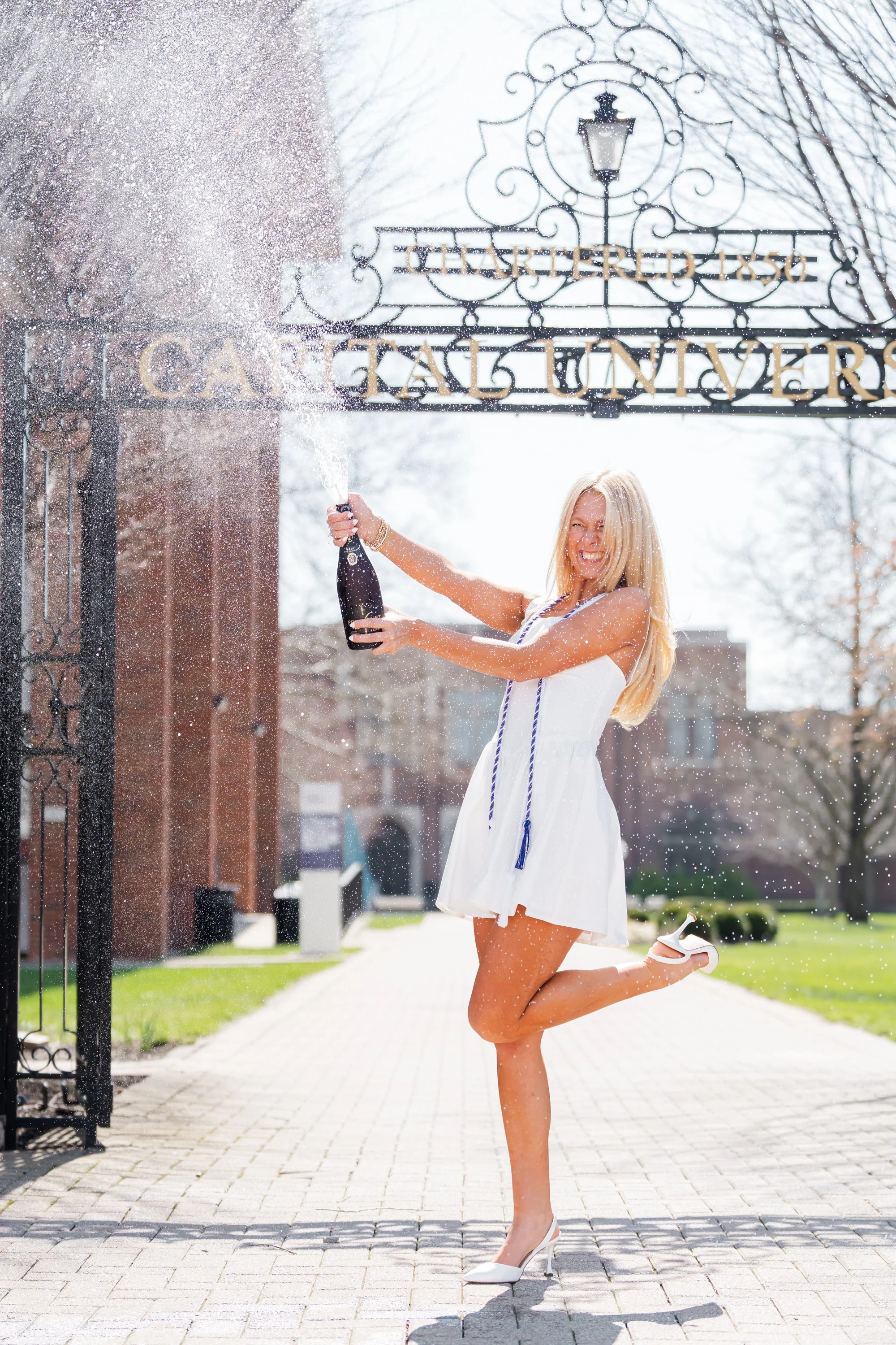 Woman in white dress celebrating graduation at Princeton University with champagne, joyfully popping the bottle, standing on campus with Princeton gates and trees in the background.