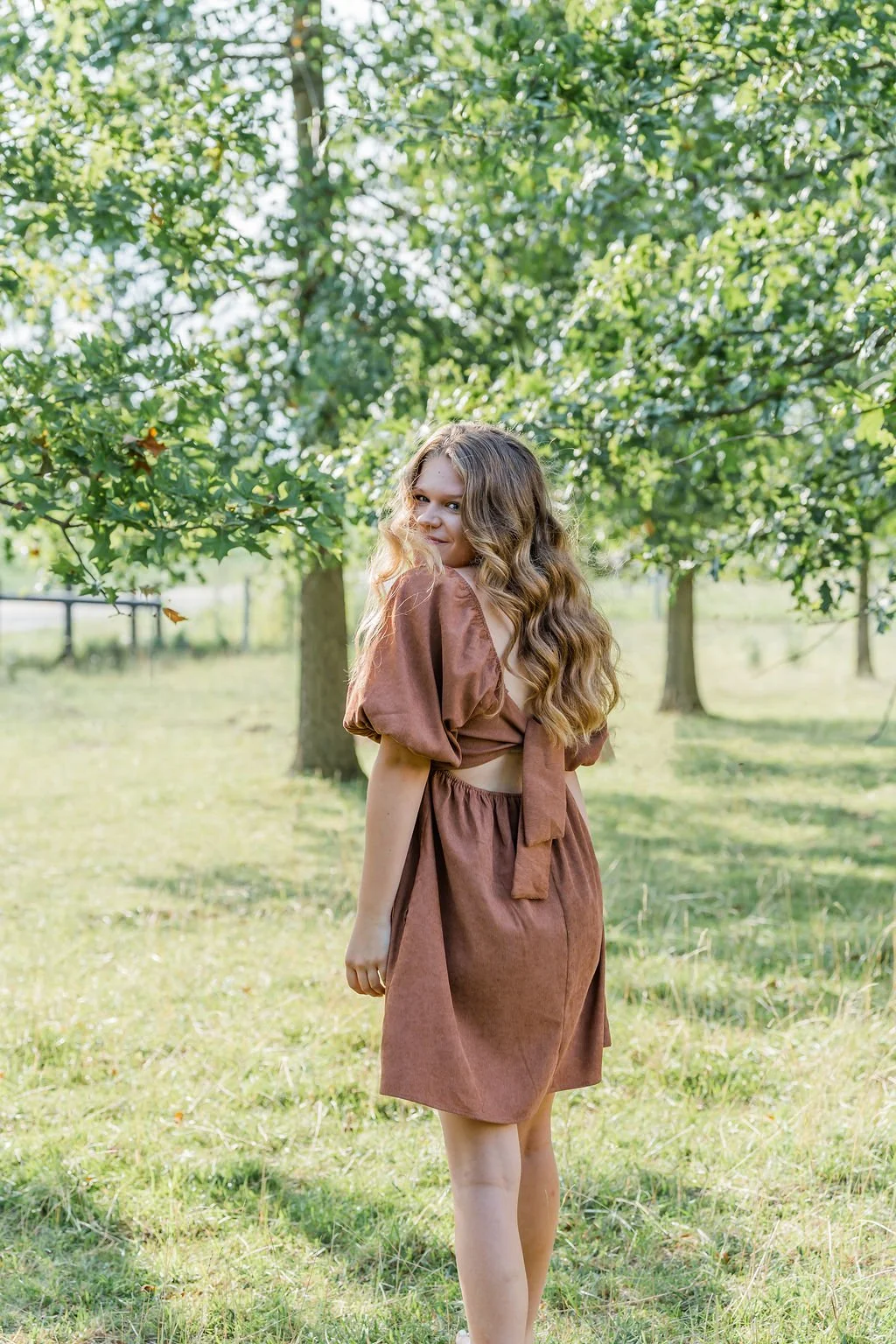 A young girl with long, wavy, light brown hair standing in a grassy park area surrounded by trees, wearing a brown two-piece outfit with puffed sleeves and a tied back top, smiling and looking over her shoulder.