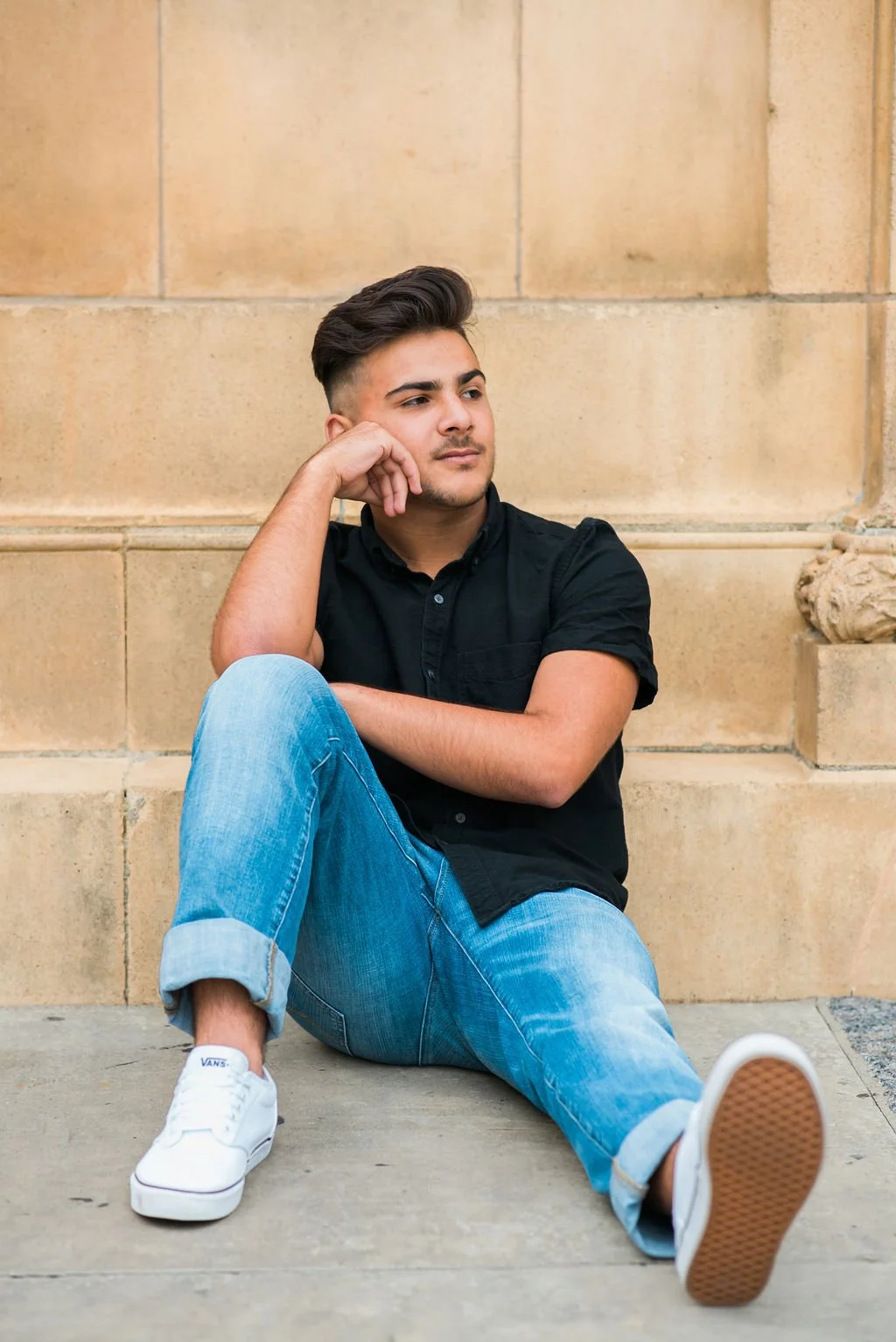 A young man sitting on the sidewalk, leaning against a stone wall, wearing a black shirt, light blue jeans, and white sneakers, with a contemplative expression.