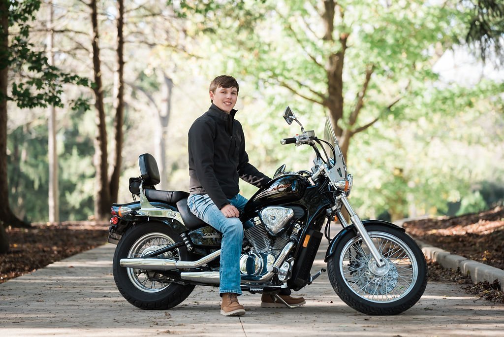 A young man sitting on a black motorcycle on a paved path in a park with trees in the background.