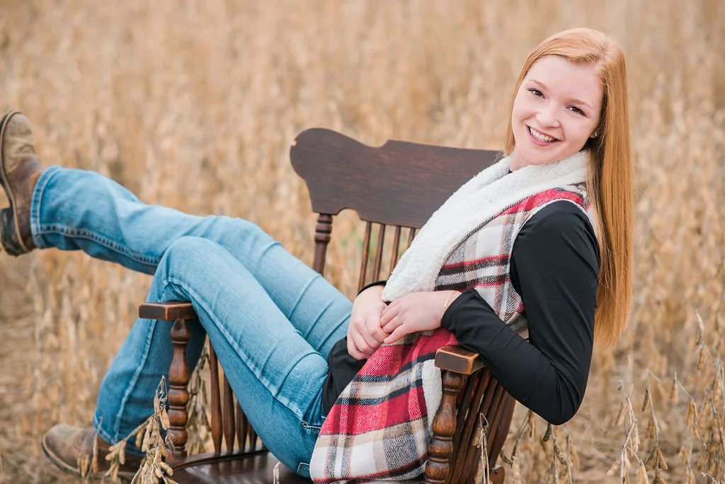 A young woman with red hair smiling and relaxing on a wooden chair outdoors in a field of tall, dried plants.