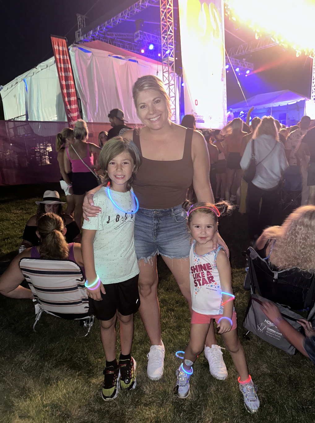 Two young girls and a woman smiling at a nighttime outdoor concert or festival. The girls are wearing glow necklaces and bracelets, and the woman is standing in the middle with her arms around them. The background shows a stage with bright lights and a crowd of people.