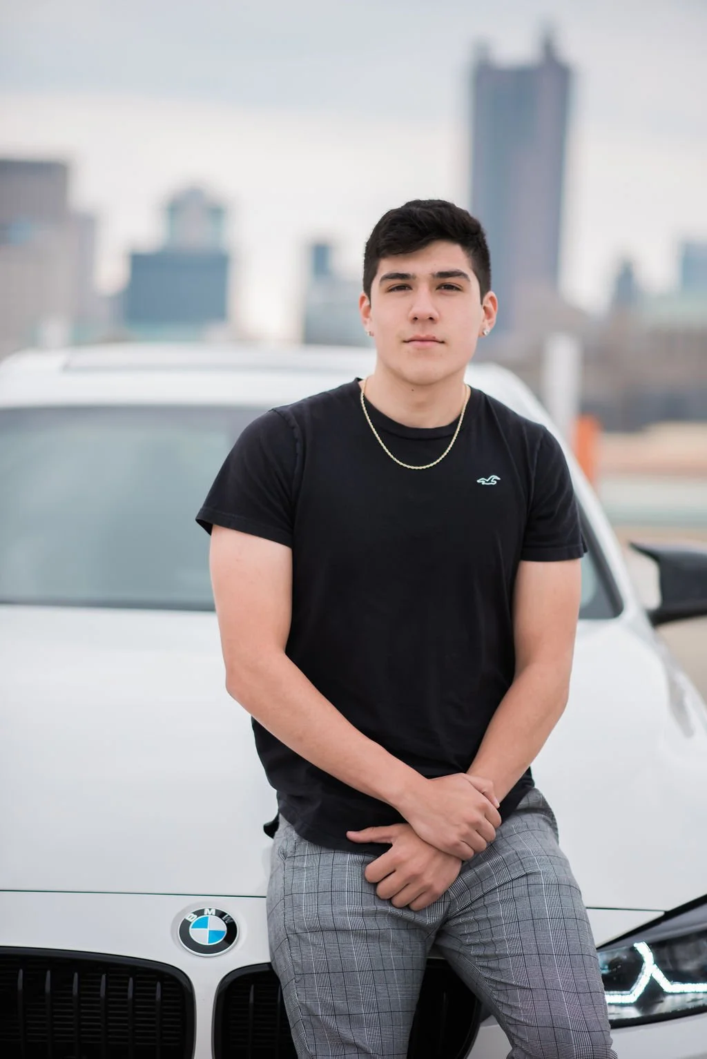 Young man with dark hair, wearing a black T-shirt and gray plaid pants, standing in front of a white BMW car in an urban setting with skyscrapers in the background.