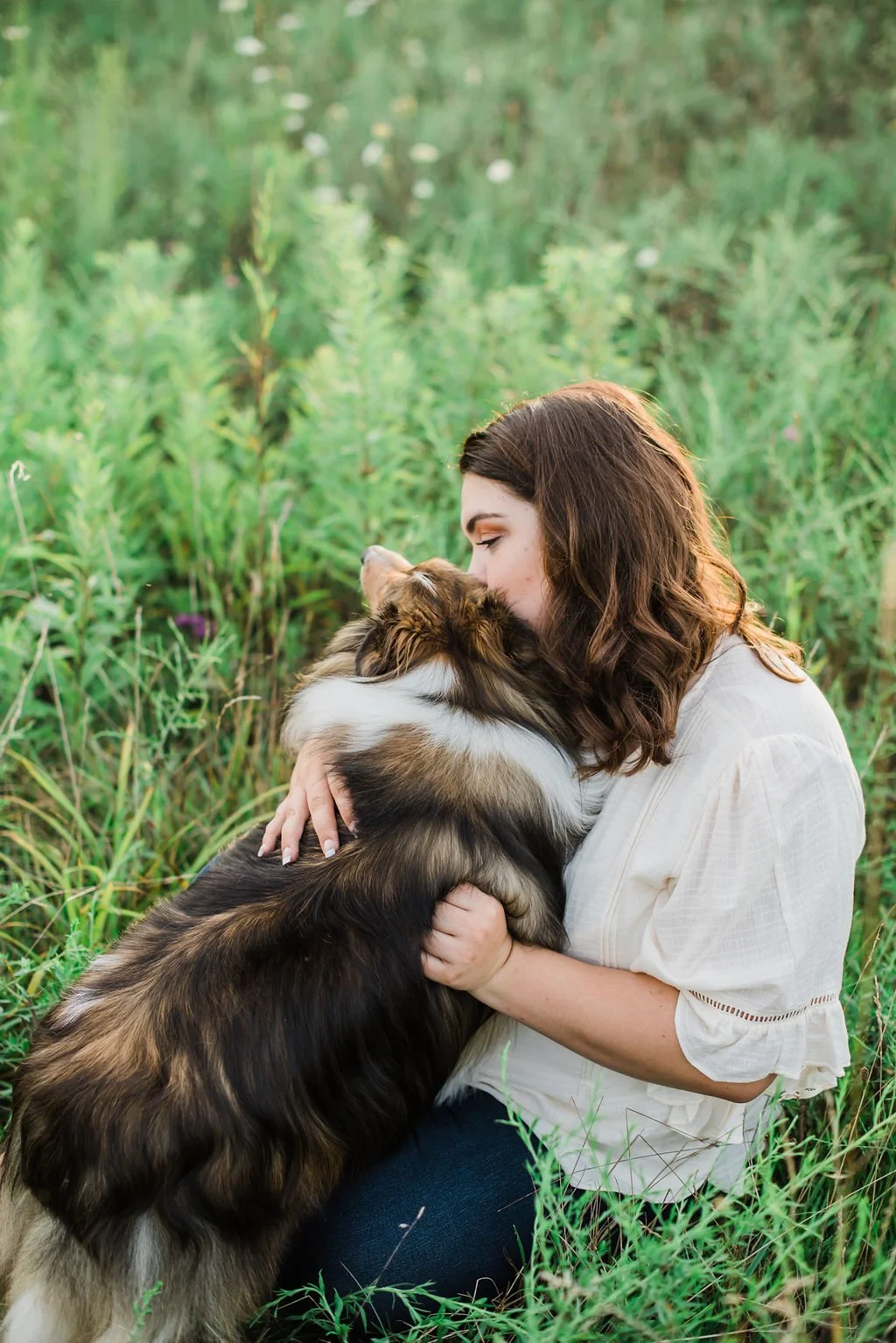 A woman with brown hair hugging and kissing a large fluffy Australian Shepherd dog in a green grassy field.