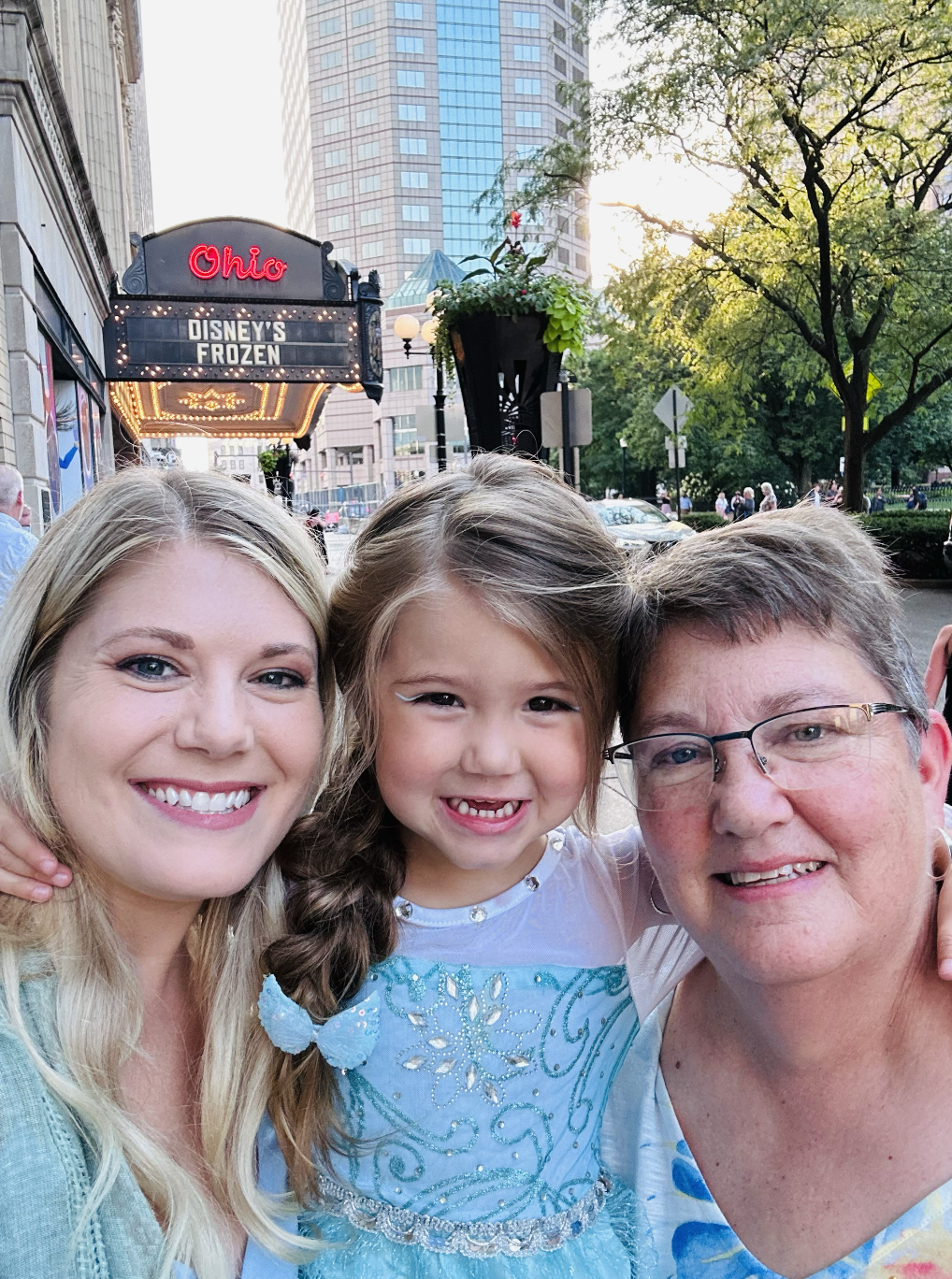 A family of three taking a selfie outside a theater with a marquee that reads Disney's Frozen. There are tall buildings and trees in the background.