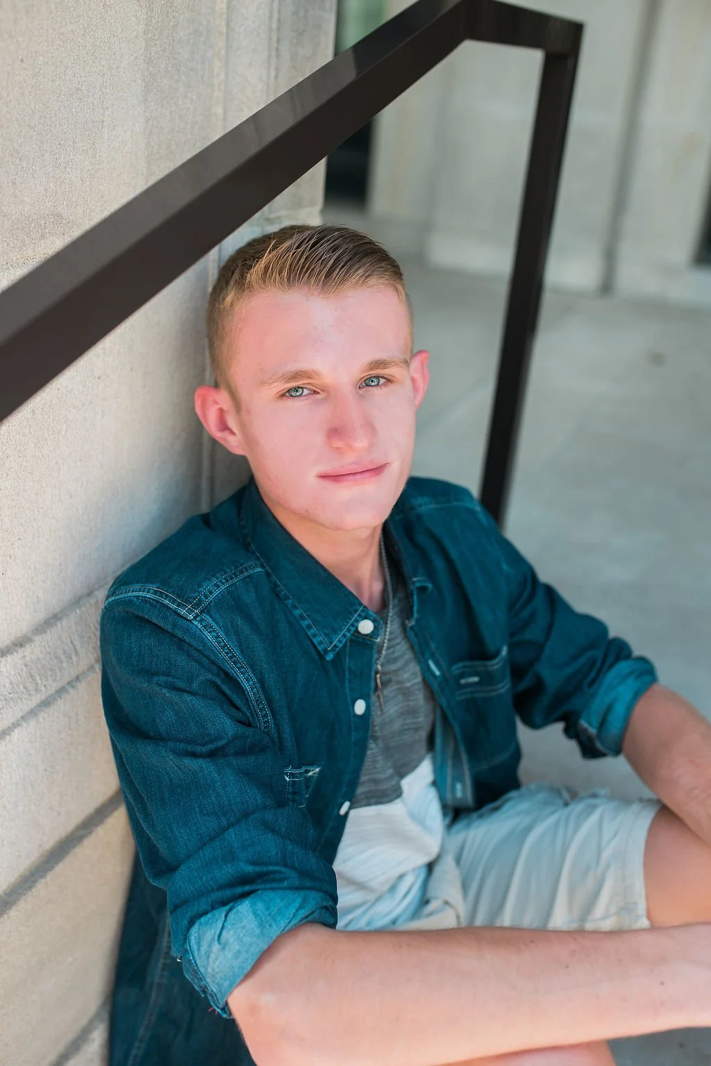 Young man with blonde hair and blue eyes sitting against a wall, wearing a denim jacket and shorts.