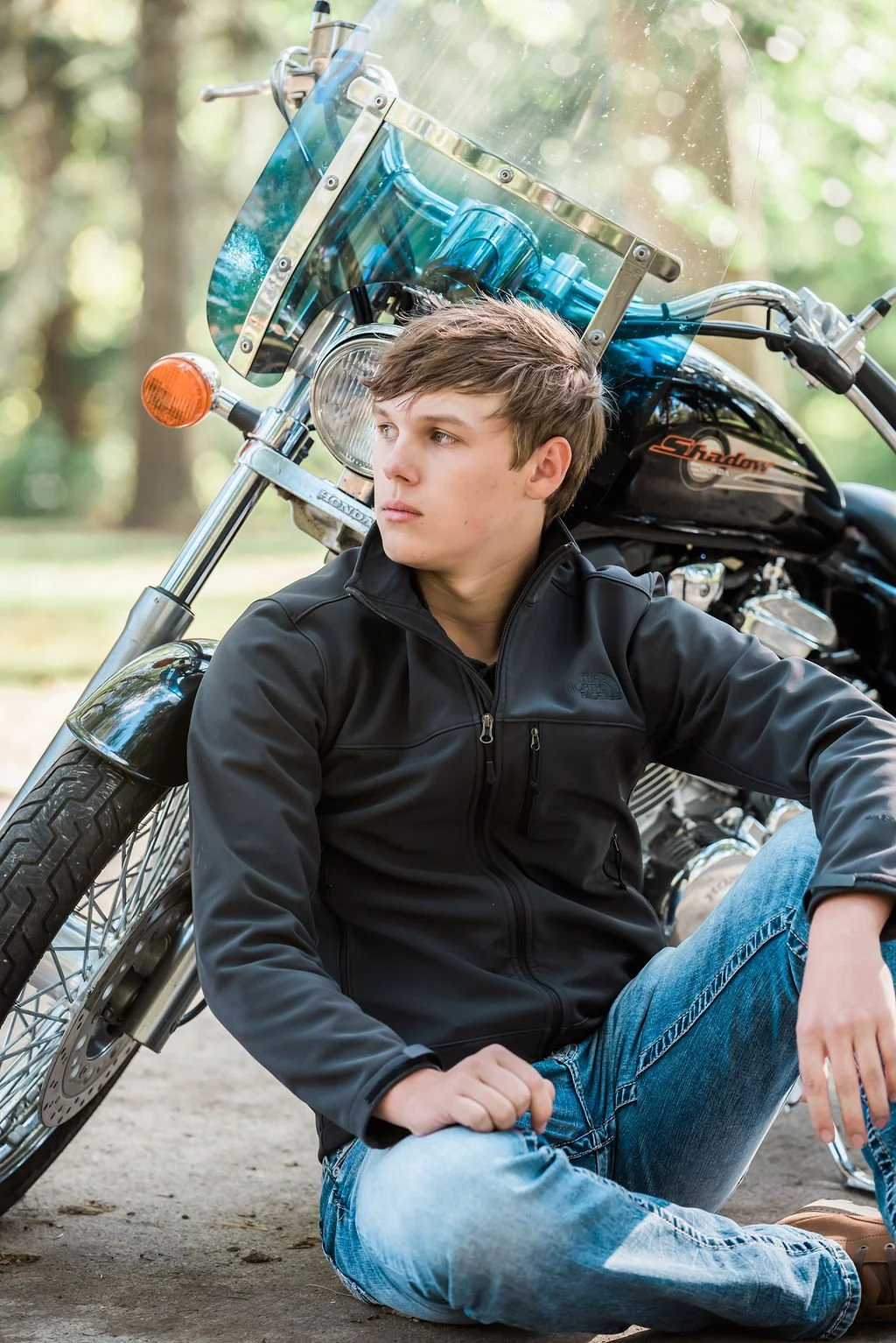A young man sitting on the ground next to a black motorcycle with a windscreen, in a forested area, wearing a black jacket and blue jeans.