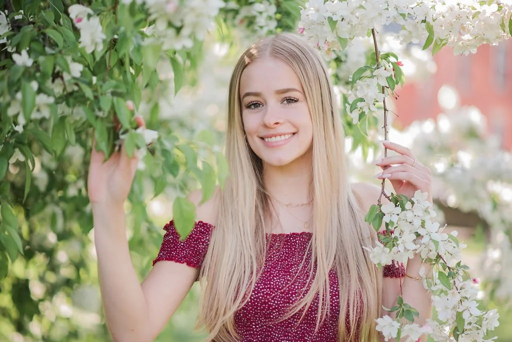 A young woman with long blonde hair smiling in a garden surrounded by blooming white flowers on green trees.