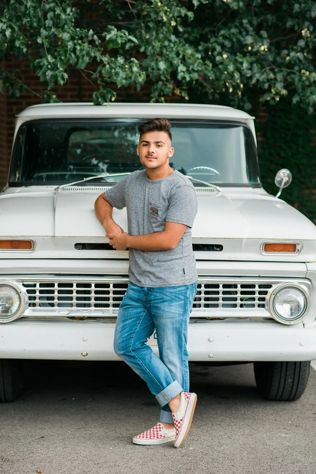 Young man standing in front of a vintage white van, leaning on its front bumper with one foot crossed over the other, wearing a gray T-shirt, blue jeans, and red checkered slip-on shoes, with green leafy trees in the background.