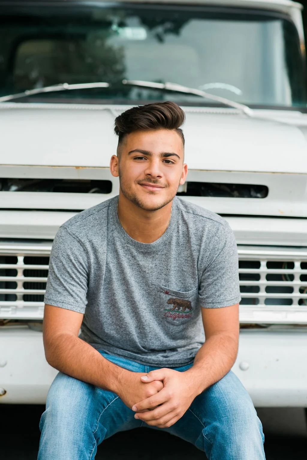 A young man with dark hair and a slight smile, wearing a gray t-shirt with a California bear logo, sitting in front of a white vintage truck.