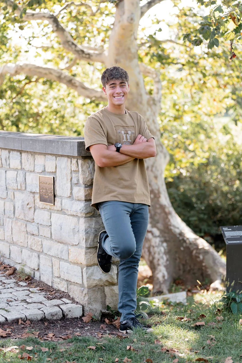A young man with curly hair in a tan t-shirt and blue jeans, smiling with arms crossed, leaning on a stone wall outdoors with trees in the background.