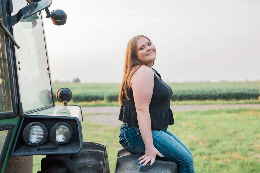 A young woman with red hair and a black sleeveless top sitting on a tractor tire in a field, smiling at the camera.