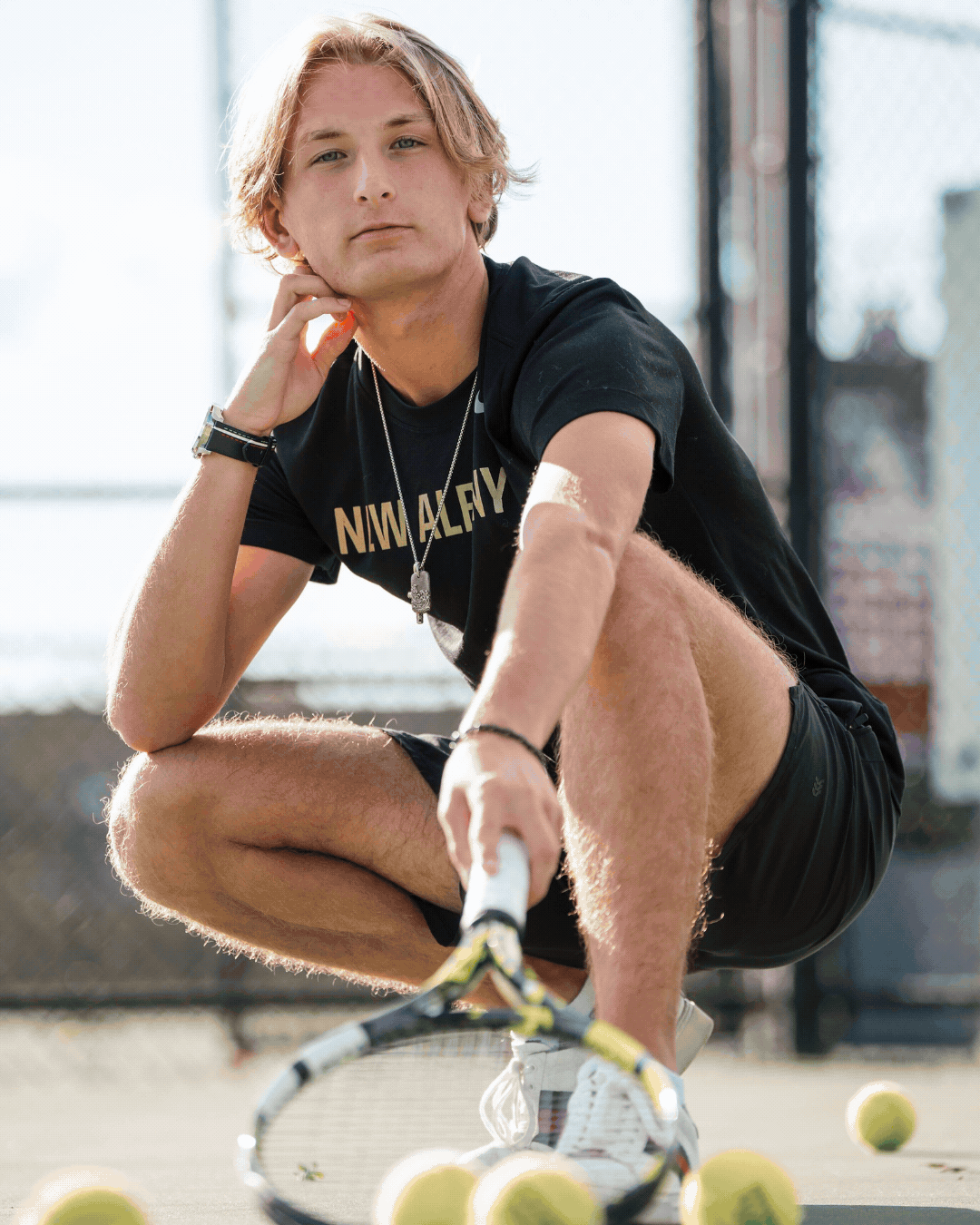 A young man crouches on a tennis court holding a tennis racket, wearing a black t-shirt and shorts, with tennis balls scattered around him.