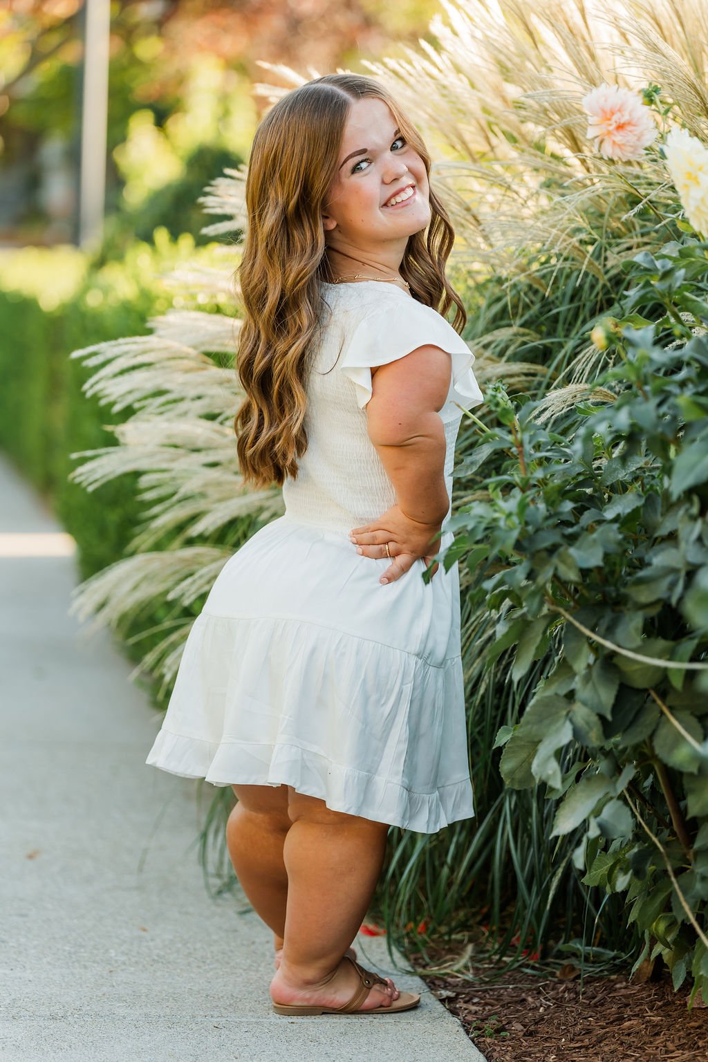 A young woman with long wavy hair wearing a white dress and sandals standing on a sidewalk beside a lush green hedge and tall ornamental grasses with pink and yellow flowers, smiling and looking at the camera in sunlight.