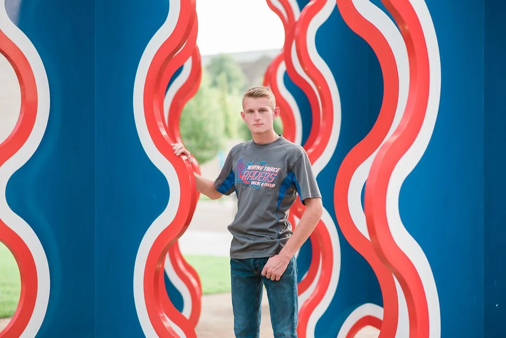 Young man in a gray T-shirt and jeans standing among wavy, colorful blue, red, white, and yellow sculptures at an outdoor art installation.