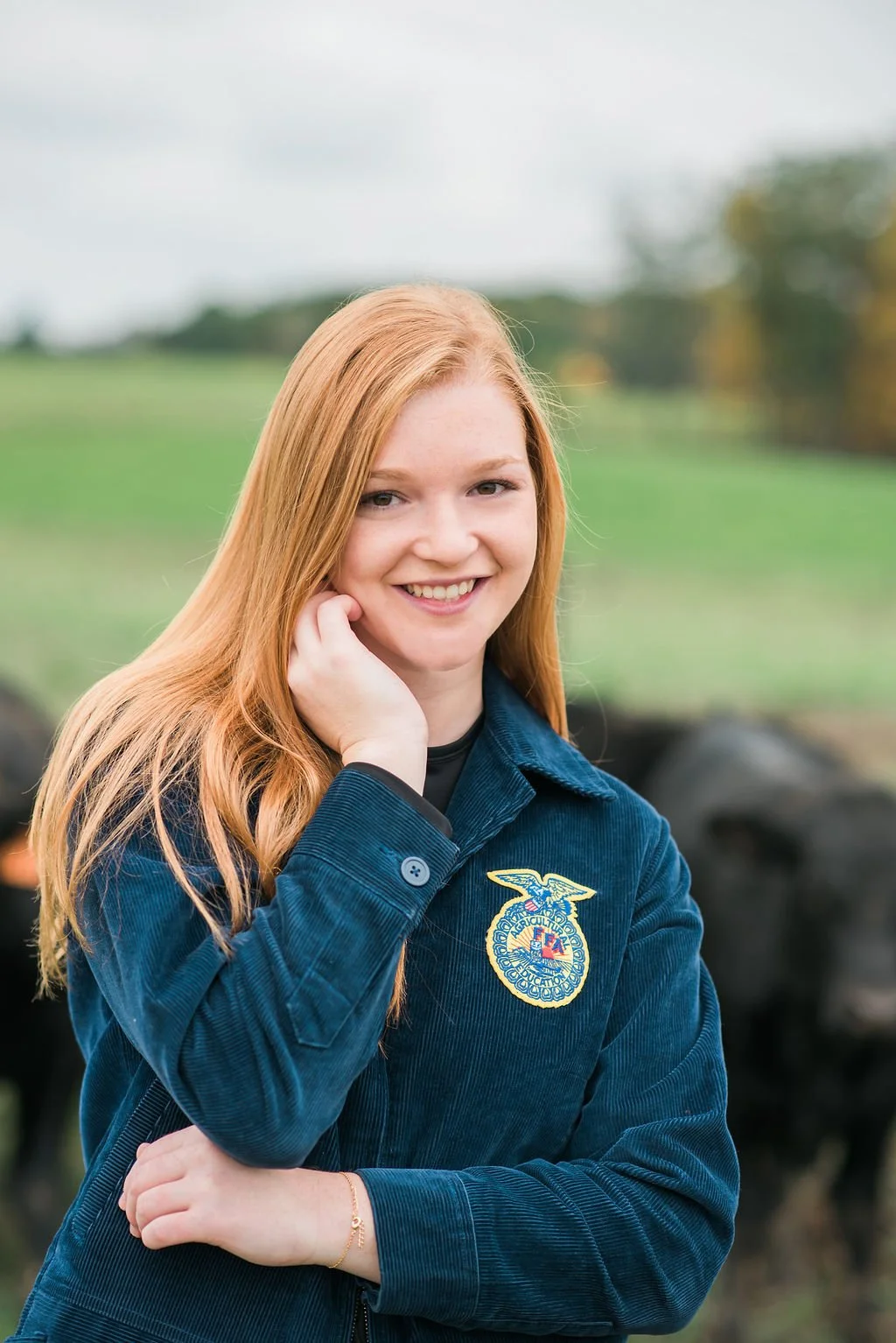 Young woman with red hair smiling outdoors, wearing a blue FFA jacket with the FFA emblem.