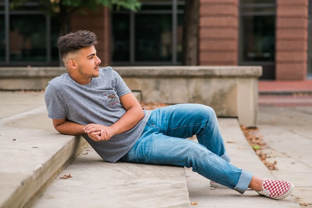 Young man with short dark hair and a beard, wearing a gray T-shirt and blue jeans, sitting on a concrete step outside near a building, looking to the left.