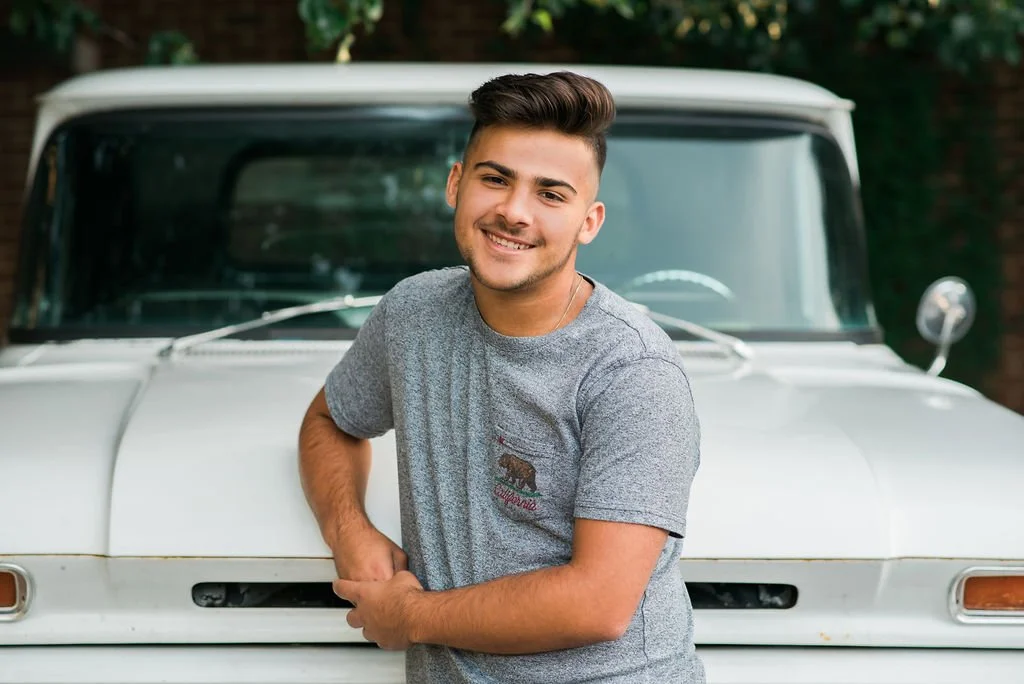 A smiling young man with short dark hair in a gray T-shirt leaning against the front of a vintage white truck outdoors.