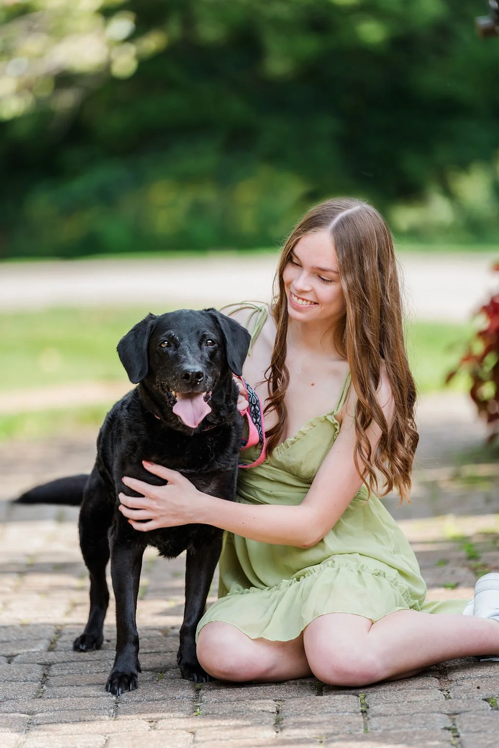 A young woman with long, wavy brown hair sitting on brick pavement, smiling while holding a happy black dog with a pink collar outdoors in a park, with green trees in the background.