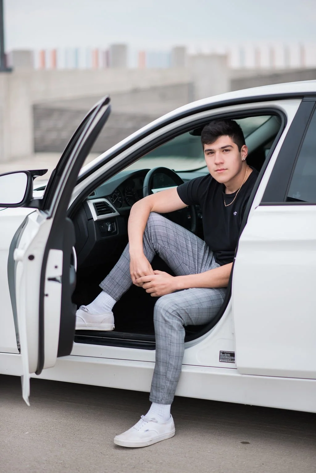 A young man with dark hair and light skin sitting in the driver's seat of a white car, looking at the camera, wearing a black t-shirt, plaid pants, white sneakers, and jewelry, with the door open.