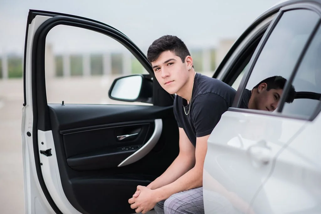 A young man sitting in the open passenger side of a white car, looking at the camera with a neutral expression.