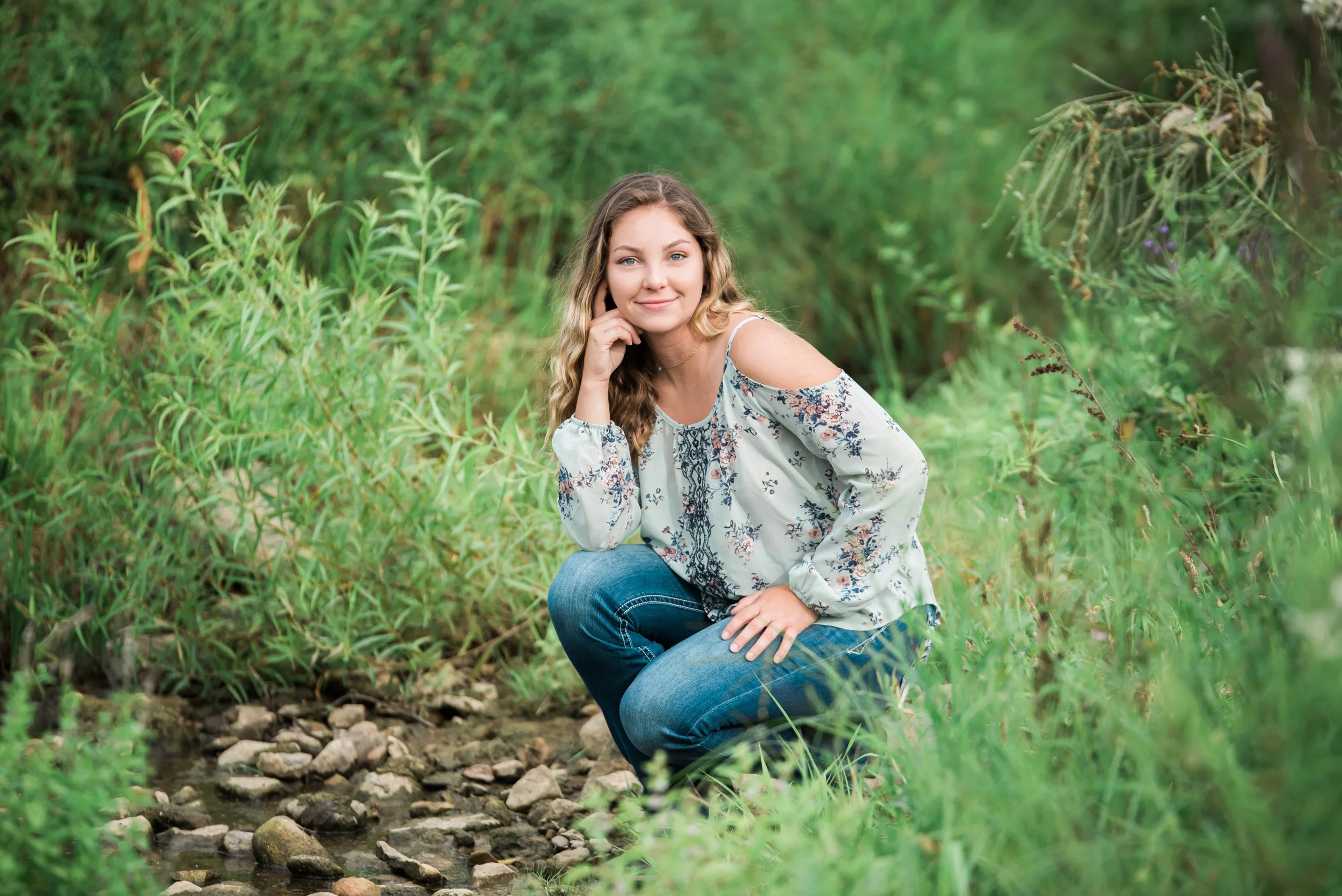A young woman with wavy hair squatting beside a small creek in a lush green forest, wearing a floral off-shoulder blouse and jeans, smiling at the camera.