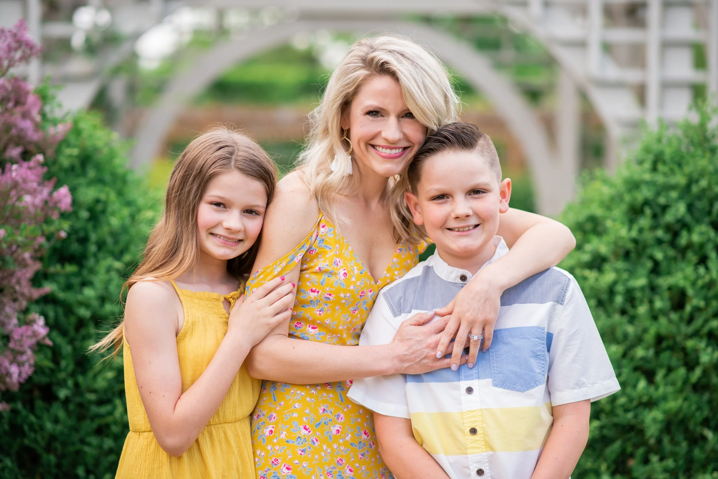 A woman with two children outside, smiling and embracing each other, in front of greenery and a garden arch.