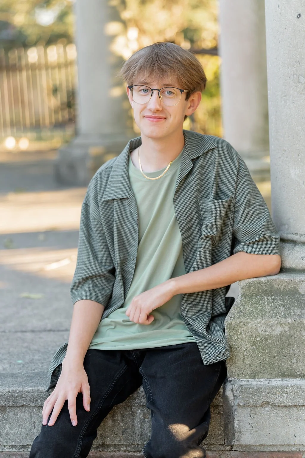 A young man with brown hair, glasses, and fair skin sitting outdoors against a concrete structure, wearing a green t-shirt, a green checkered shirt, and black pants, smiling at the camera.