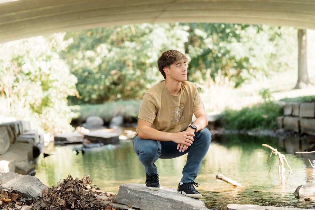 Young man kneeling on a rock by a creek under a bridge, looking thoughtful, surrounded by greenery.