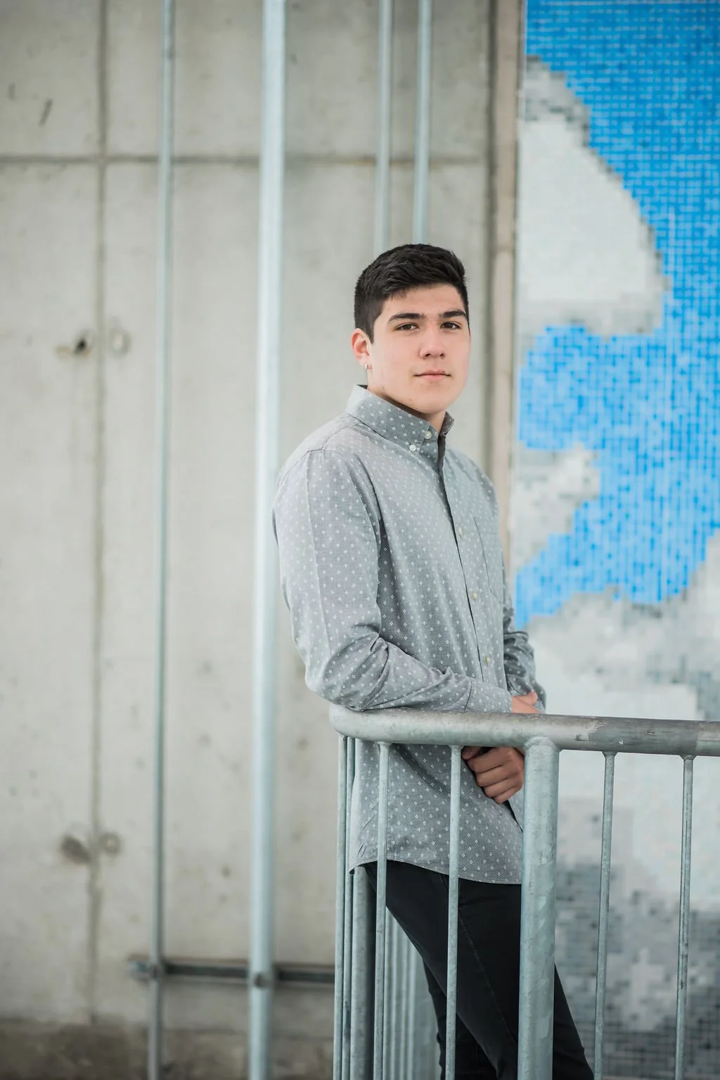 Young man with dark hair wearing a gray polka dot shirt leaning against a metal rail in an industrial-style setting with concrete walls and a blue mosaic corner.