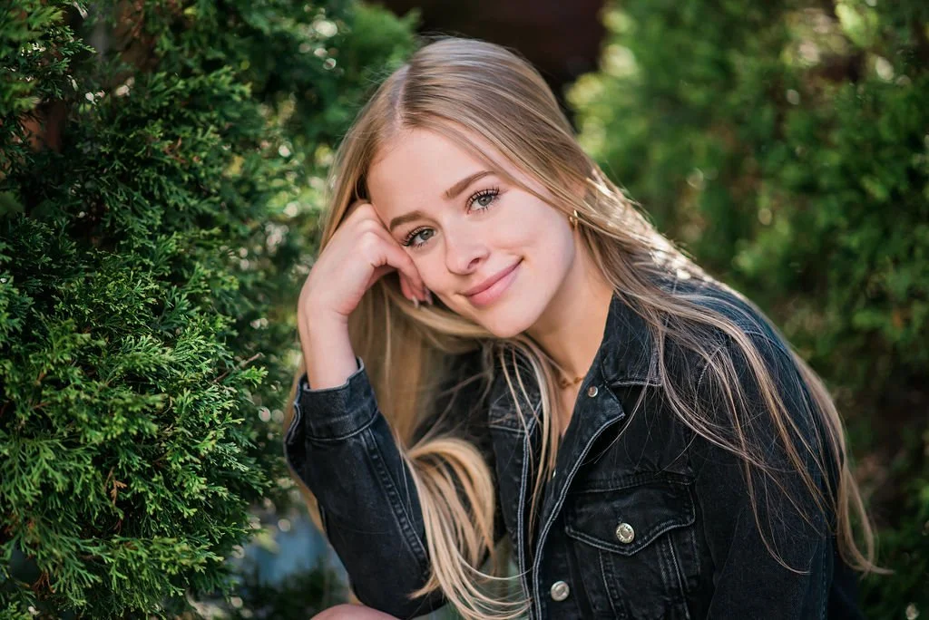 A young woman with long blonde hair, smiling, resting her head on her hand, outdoors near green bushes.