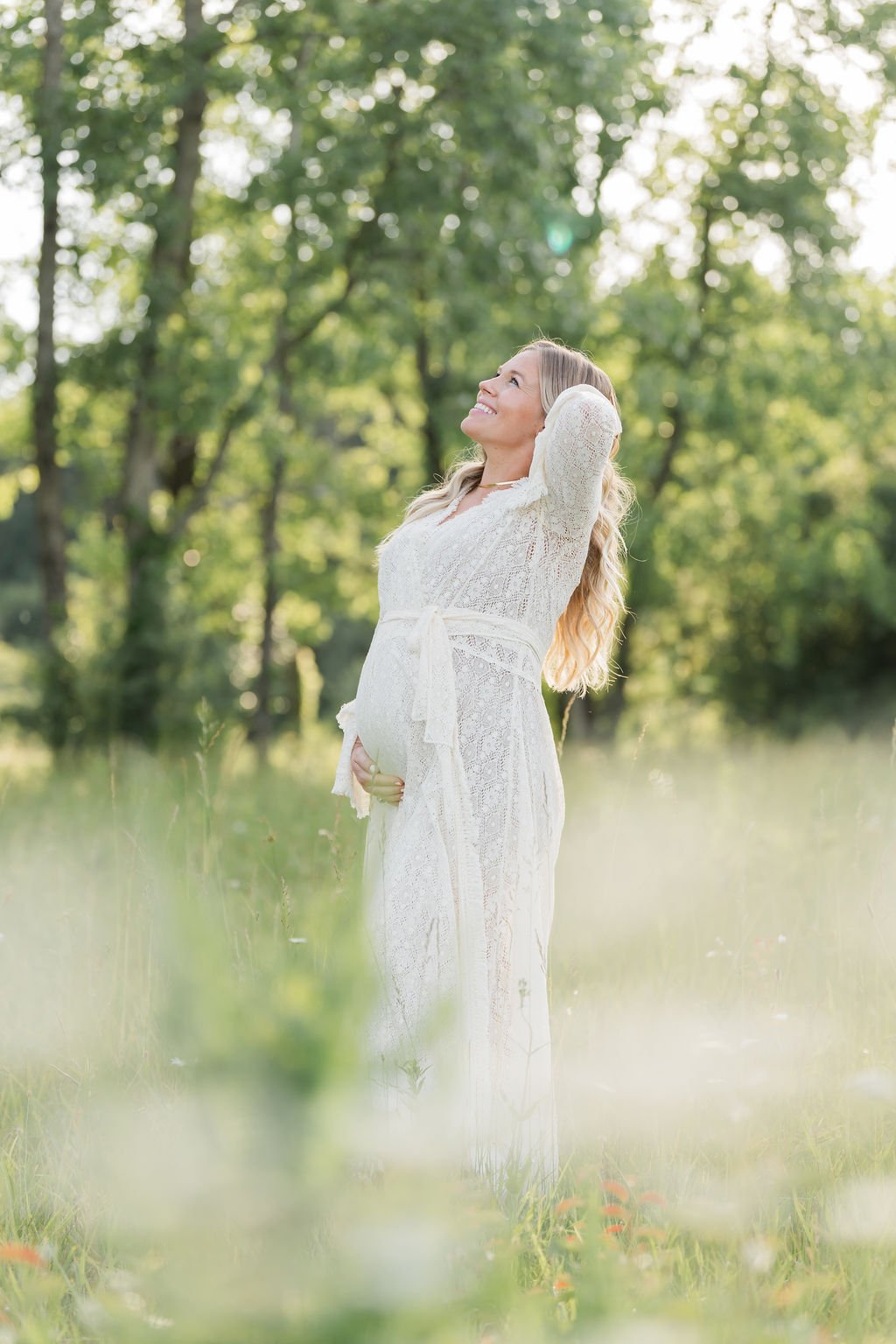Pregnant woman in a white lace dress standing in a lush green field with trees in the background, smiling and looking upwards with sunlight filtering through the trees.