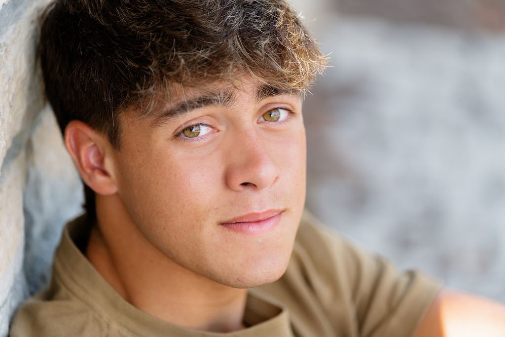 Close-up of a young man with light brown hair and hazel eyes, leaning against a stone wall and looking at the camera.