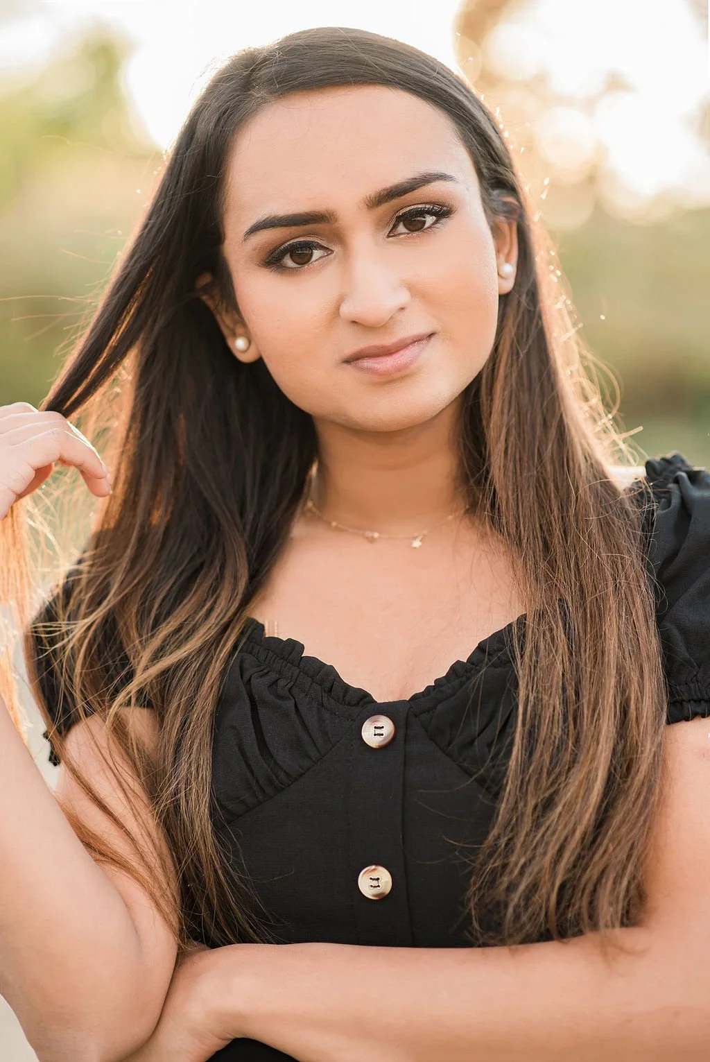 A young woman with long brown hair, wearing a black top with large buttons, pearl earrings, and a delicate necklace, stands outdoors with sunlight in the background.