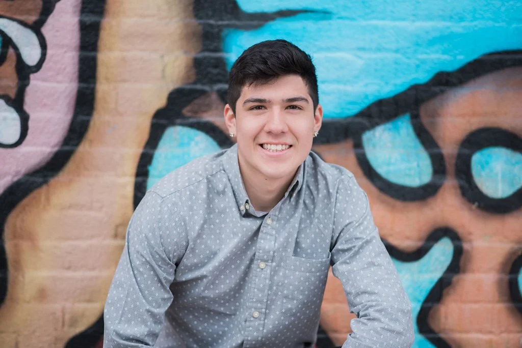 A young man with dark hair smiling, wearing a gray dotted button-up shirt, sitting in front of a colorful graffiti wall.