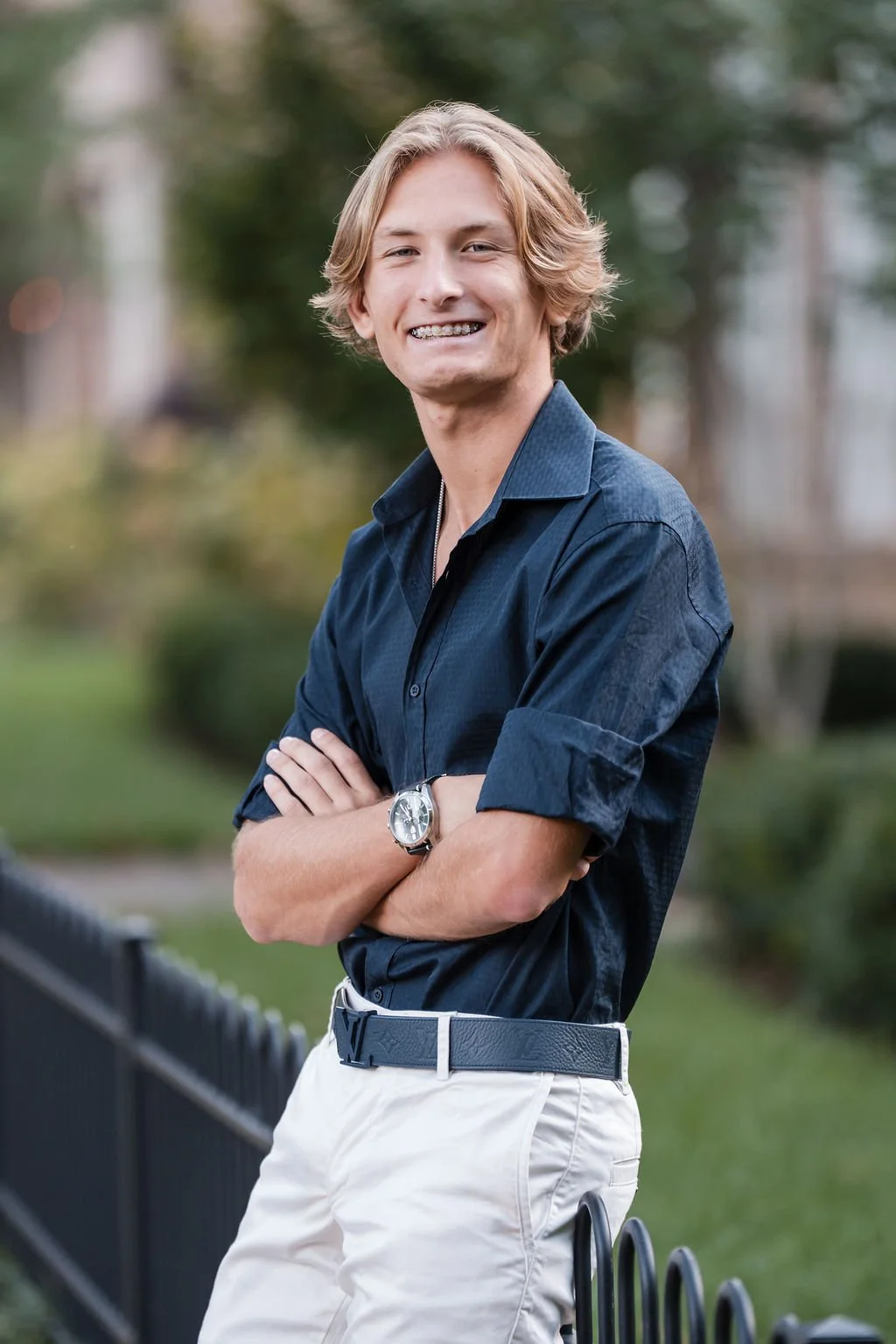 A young man with blond hair, wearing a dark blue shirt and white pants, standing outdoors with his arms crossed and smiling.