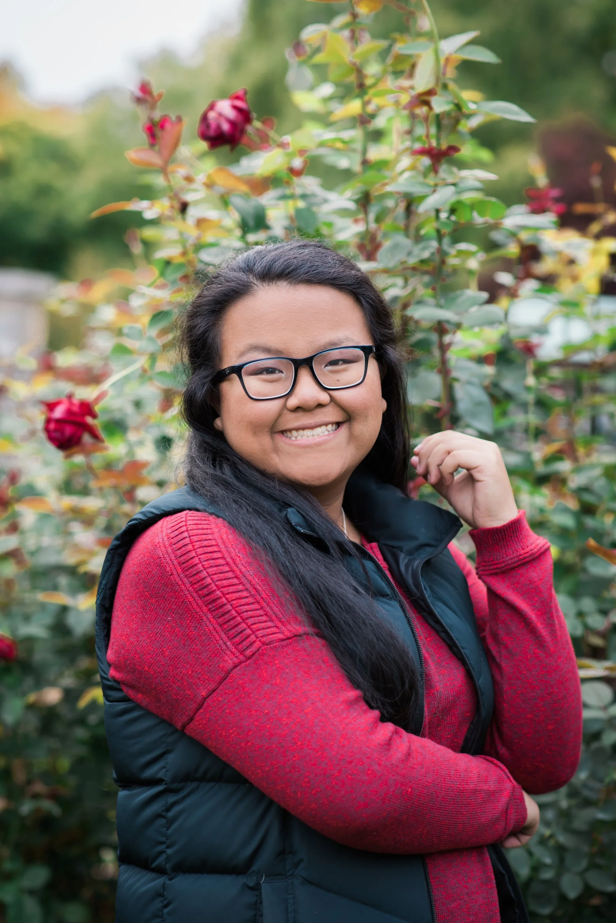 Smiling woman with glasses and long black hair, wearing a red sweater and black vest, standing outdoors in front of a background of pink roses and green foliage.