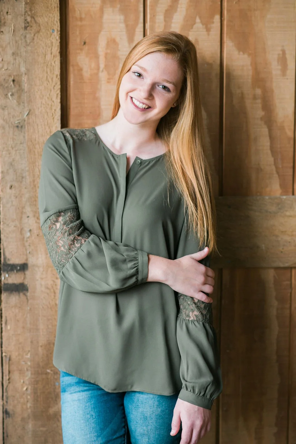 A young woman with long red hair smiling and standing with arms crossed, leaning against a wooden wall, wearing an olive green blouse with lace detailing on the sleeves and blue jeans.
